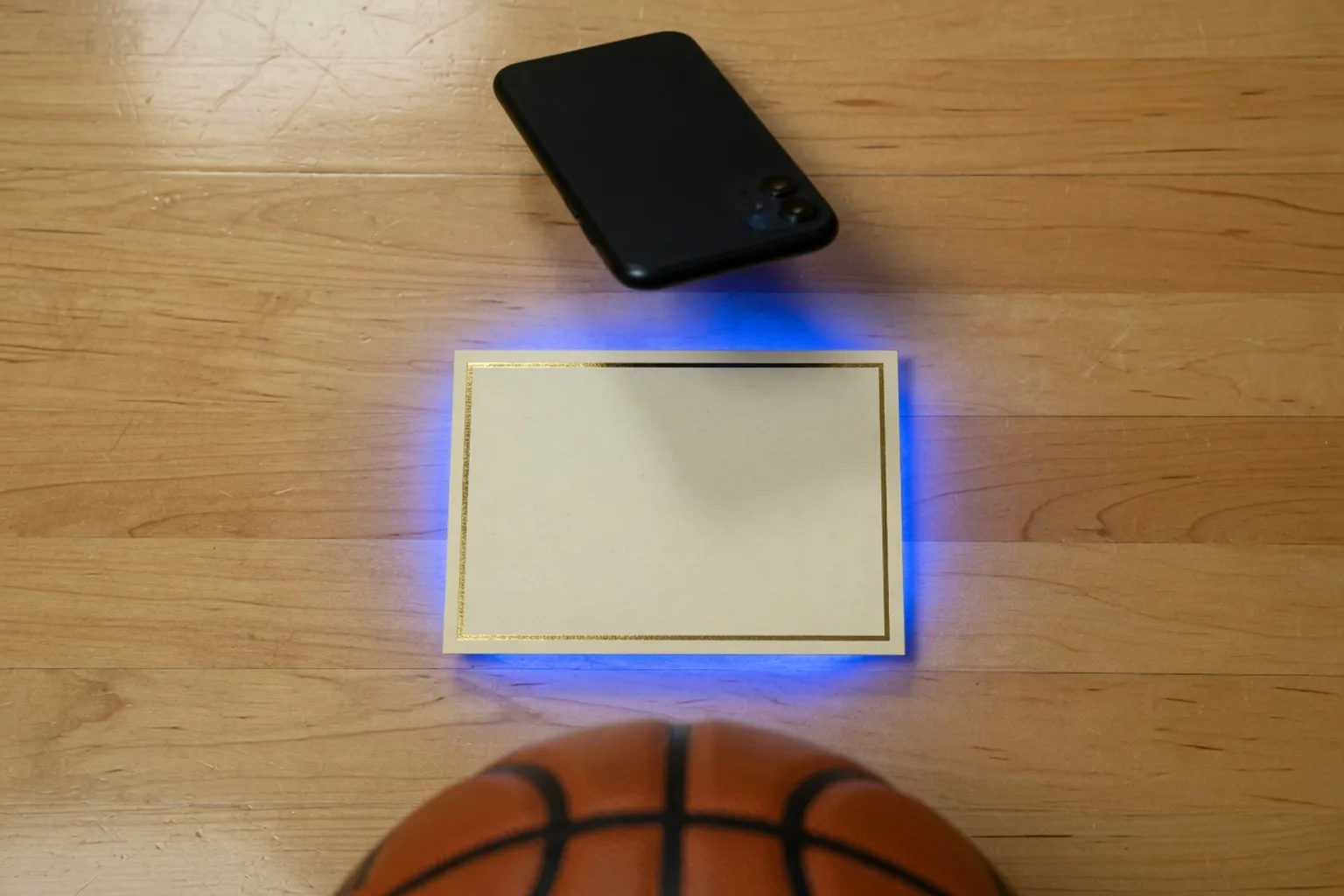Black phone hovering over a gold-bordered blank card on hardwood, basketball at the bottom of the frame
